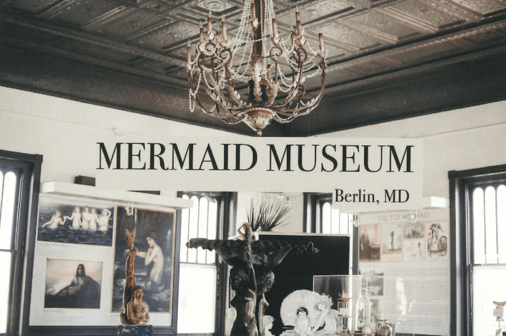 Interior view of the Mermaid Museum in Berlin, Maryland. A chandelier hangs from a decorative ceiling above mermaid-themed exhibits and displays. The text 'MERMAID MUSEUM' and 'Berlin, MD' is prominently featured in the center of the image.