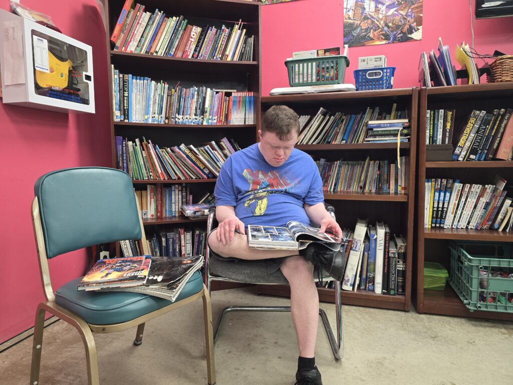 A person sits in a library with pink walls, reading a book. They are wearing a blue graphic t-shirt and black shorts. Behind them are bookshelves filled with books. To their left is an empty green chair with several magazines or comic books on it.