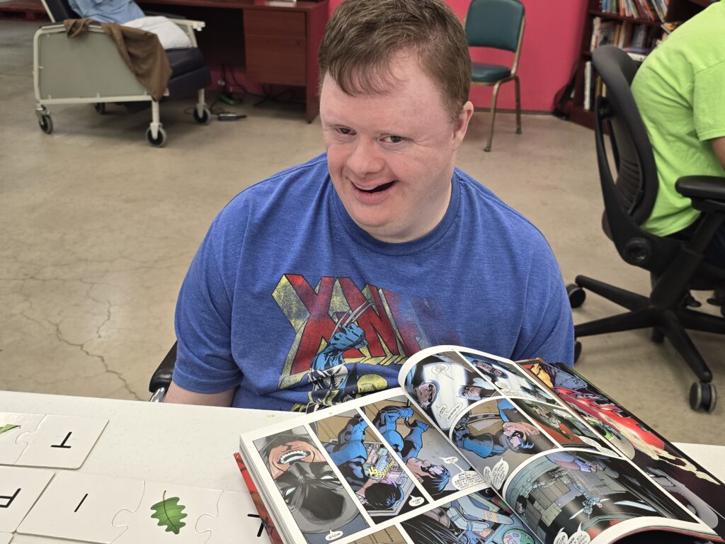 A person wearing a blue X-Men t-shirt is sitting at a table, reading a comic book. The table is covered with puzzle pieces featuring letters and leaf illustrations. In the background, another person in a green shirt sits in an office chair near bookshelves against pink walls.