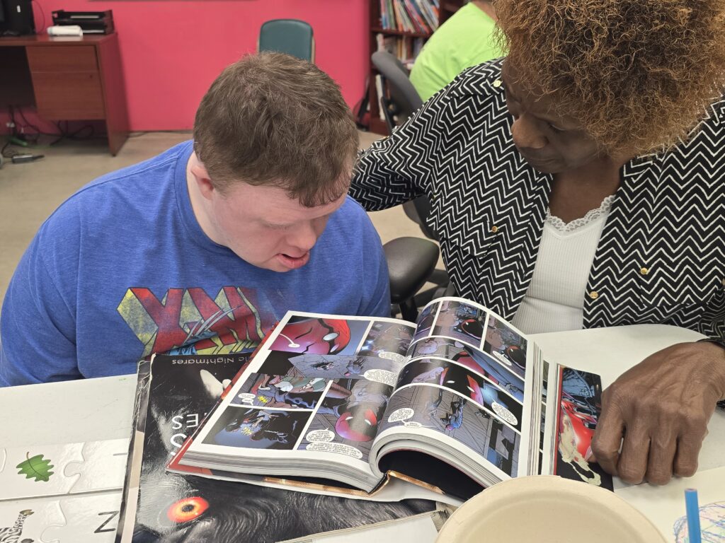 Two people are sitting at a table, looking at an open comic book. The person on the left is wearing a blue X-Men t-shirt, and the person on the right is wearing a black and white zigzag patterned jacket over a white shirt. The table has puzzle pieces with letters and leaf illustrations. In the background, there are bookshelves against pink walls.