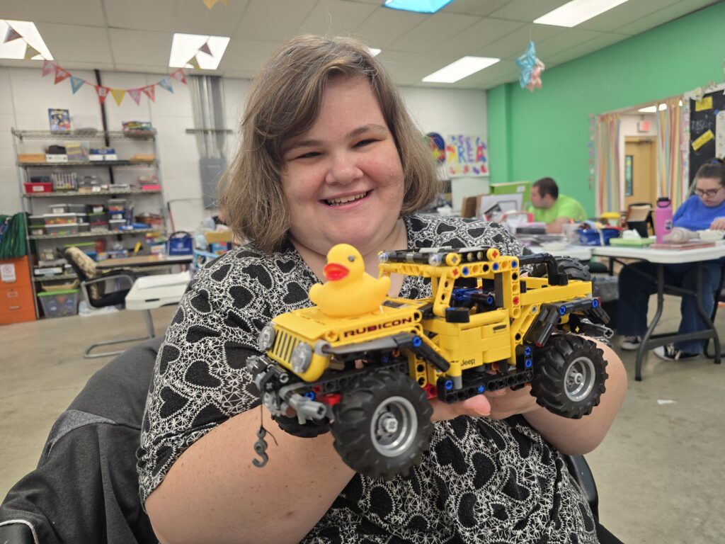 A person wearing a black and white patterned shirt holds a yellow toy truck made from Lego building blocks, with a rubber duck on top. The truck has 'Rubicon' written on its side. In the background, shelves with various items, colorful ceiling decorations, and people sitting at tables are visible in a room with green walls.