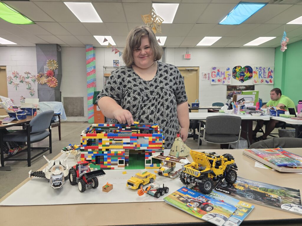 A person wearing a black and white patterned shirt stands behind a table displaying various LEGO creations, including a large multicolored structure, several vehicles, and a yellow toy truck. The room has green walls, tables, chairs, other people engaged in activities, colorful ceiling decorations, and artwork on the walls.