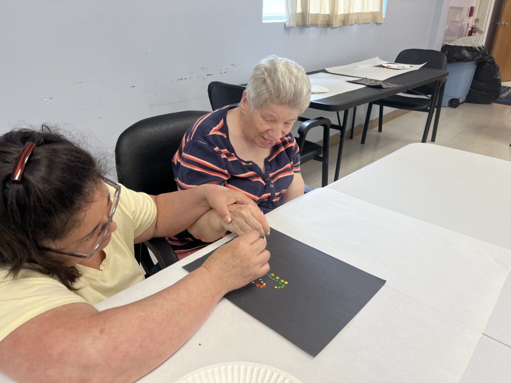 Two people are seated at a table, working on an art project. The person on the left, wearing a yellow shirt, is assisting the person on the right, who is wearing a striped shirt. They are placing small colorful objects onto a black piece of paper. The room has light blue walls, windows with curtains, and additional tables and chairs in the background.