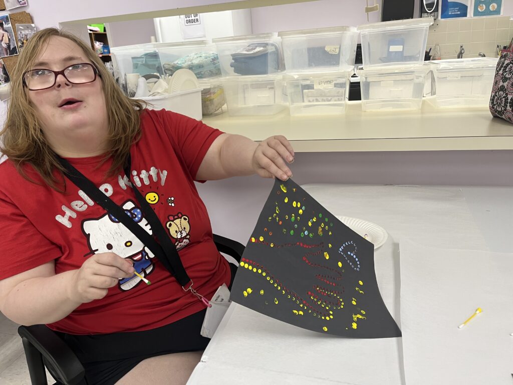 A person wearing a red Hello Kitty t-shirt sits at a white table, holding up a piece of black paper decorated with colorful dots and lines made from small objects. Behind them are shelves with clear plastic storage containers.