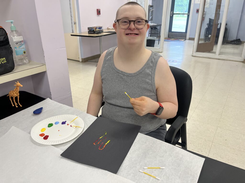 A person wearing a gray tank top sits at a table, holding a yellow cotton swab and working on an art project. In front of them is a piece of black paper with colorful paint dots and lines. A white paper plate with various paint colors and several cotton swabs are also on the table. In the background, shelves hold items including hand sanitizer and a small giraffe figurine, with doors leading to another room.