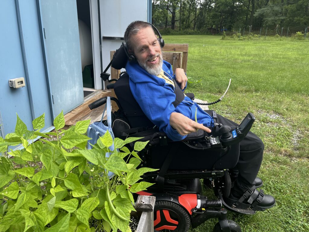 A person in a blue jacket sits in a motorized wheelchair with red accents, holding the joystick control. They are outdoors near a blue building, with green plants in the foreground and a grassy area with trees in the background.