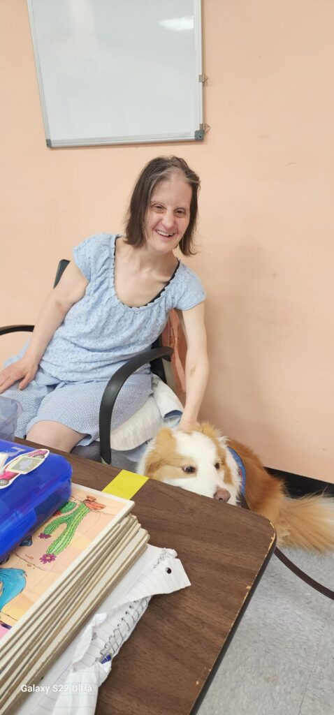 A person wearing a light blue dress sits in a chair next to a table, gently petting a white and brown dog lying on the floor. The table holds colorful drawings and papers. In the background, a light orange wall features a whiteboard.