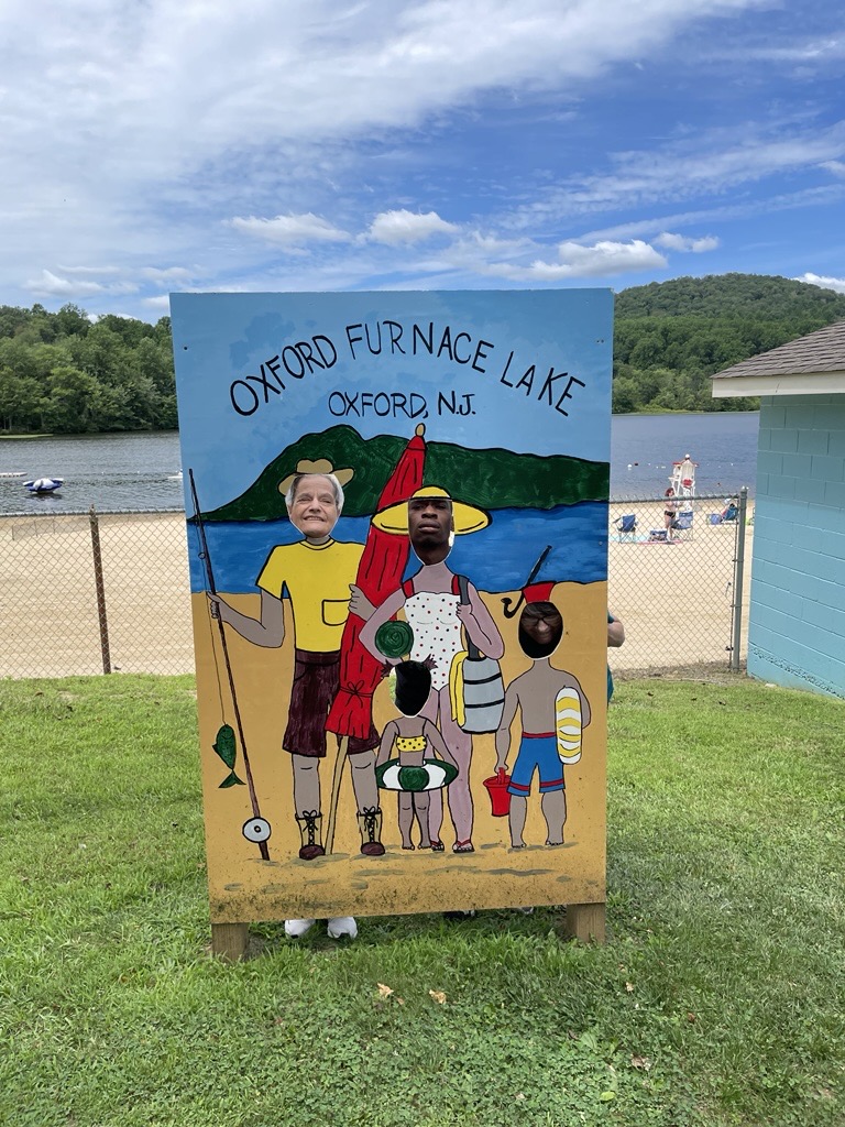 A painted cutout board at Oxford Furnace Lake in Oxford, New Jersey, shows a cartoon-style family of four on a beach with fishing gear and beach accessories. Two people pose behind the board with their faces visible through the cutouts. In the background, a lake, trees, and hills are visible under a partly cloudy sky.