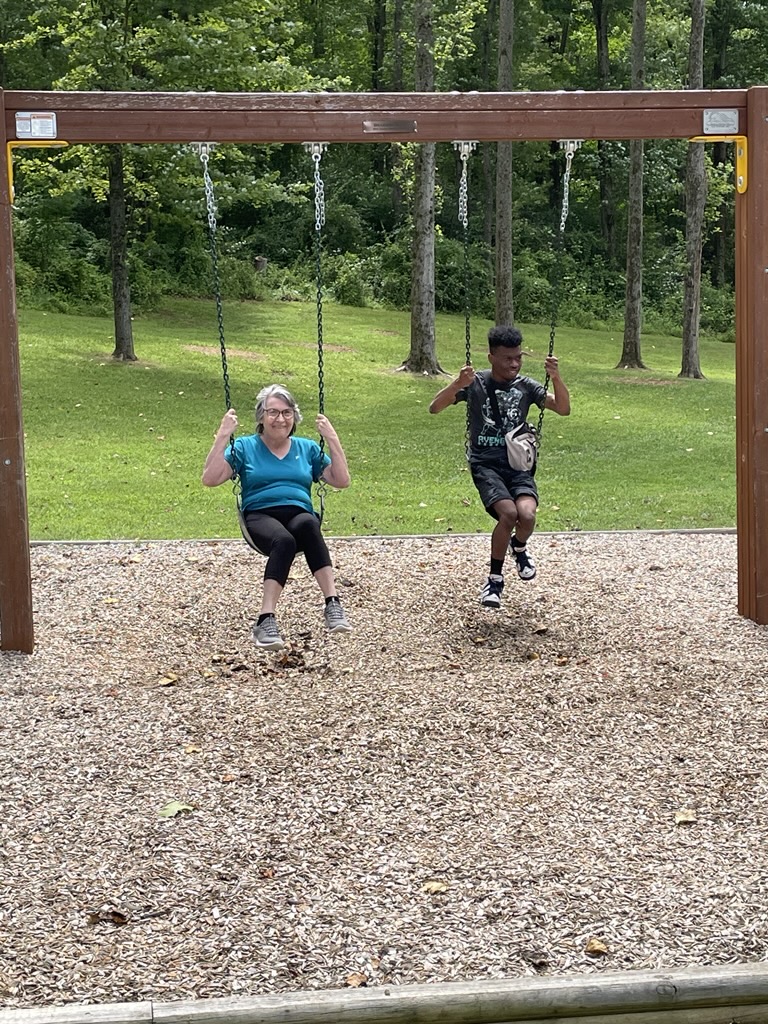 Two people are swinging on a wooden swing set in a park. The person on the left wears a teal shirt and black pants, and the person on the right wears a black t-shirt and shorts. The ground is covered with wood chips, and trees and grass are visible in the background.
