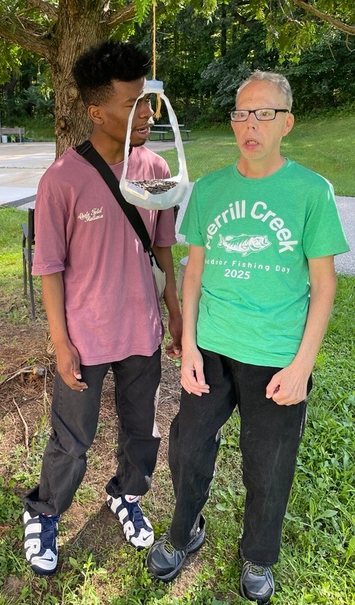 Two people stand on grass near a tree. The person on the left wears a maroon shirt and black pants, and the person on the right wears a green shirt with 'Merrill Creek Junior Fishing Day 2025' printed on it. A DIY bird feeder made from a cut open plastic milk jug is suspended from a tree branch above them. Trees and greenery are in the background.