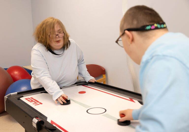 Two individuals are playing air hockey on a table with the ESPN logo. In the background, several large exercise balls and a red chair are visible against the wall.
