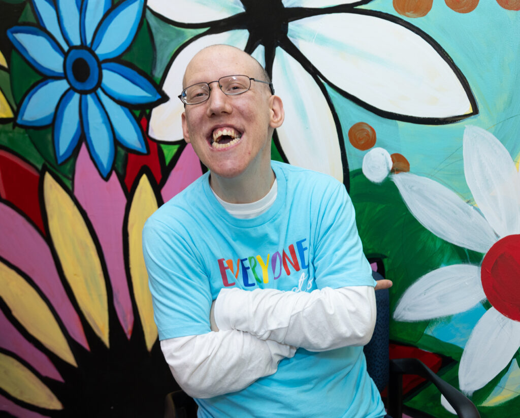 A person is standing in front of a colorful mural featuring large, vibrant flowers. The person is wearing a light blue shirt with the word "EVERYONE" written in rainbow colors and has their arms crossed.