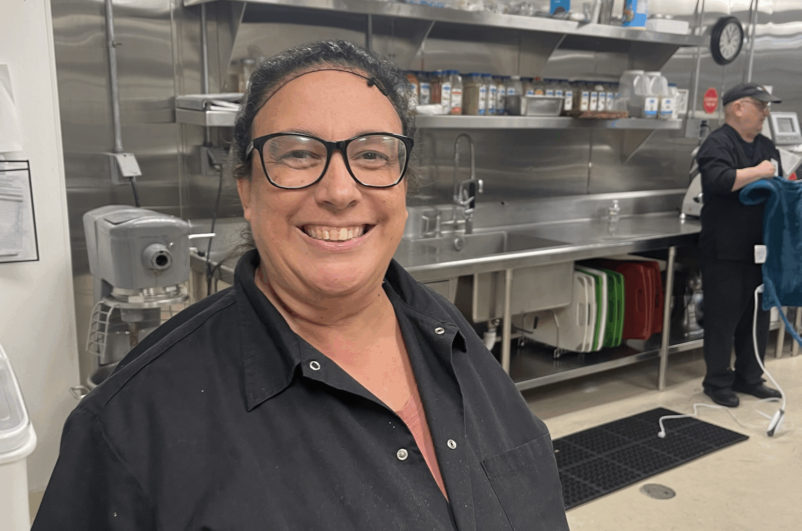 A person wearing a black shirt and glasses is smiling in a commercial kitchen. The kitchen features stainless steel appliances, shelves with various items, and another person working in the background.