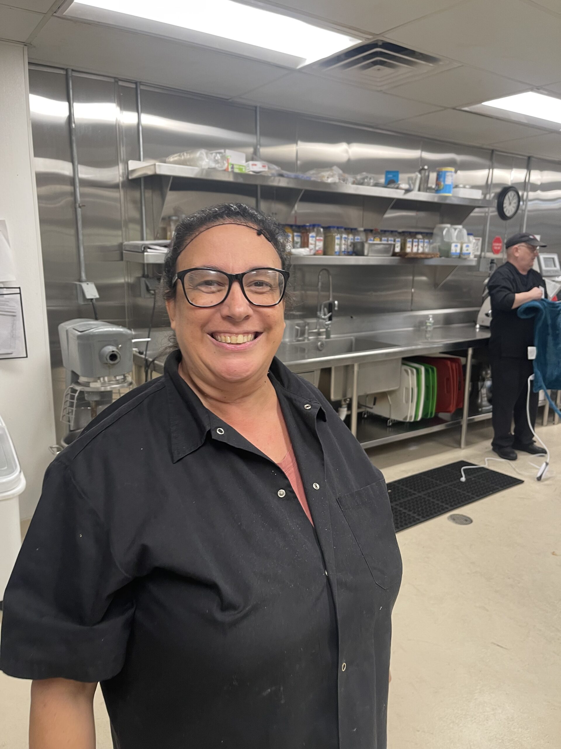 A person wearing a black shirt and glasses is smiling in a commercial kitchen. The kitchen features stainless steel appliances, shelves with various items, and another person working in the background.