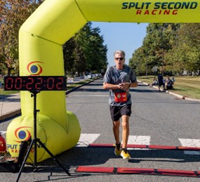 A runner crosses the finish line under a yellow inflatable arch labeled 'Split Second Racing' with a digital timer displaying 00:22:02. Spectators sit on the grass in the background near trees on a sunny day.
