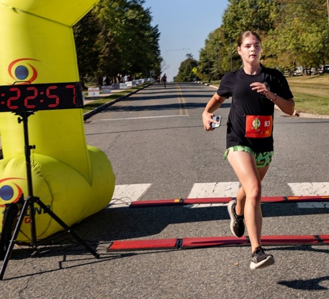 A runner wearing a black shirt, green patterned shorts, and a red bib numbered 83 crosses the finish line under a yellow inflatable arch with a digital timer showing 22:57. The race takes place on a tree-lined paved road with spectators and other runners visible in the background.