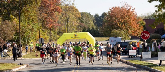 Runners approach the camera on a tree-lined street during an autumn 5K race. A yellow inflatable arch in the background reads 'Split Second Racing.' Spectators line the street, and fall foliage in red, orange, and yellow colors surrounds the scene. A vehicle with a 'Warren' banner is visible on the right.