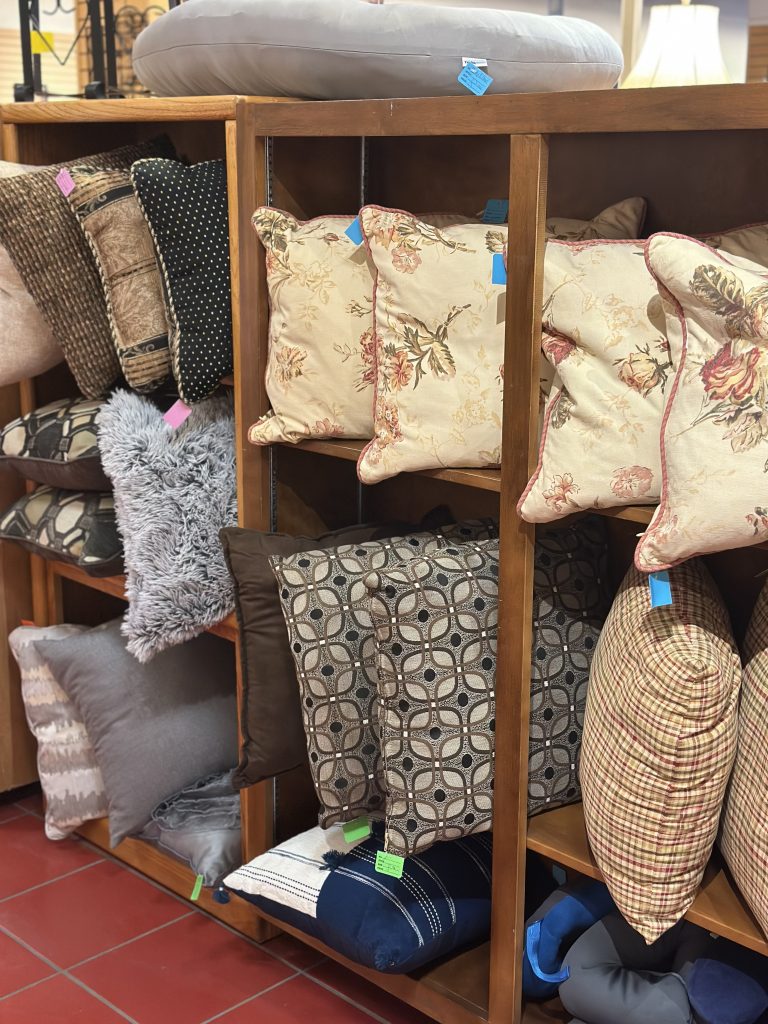 Shelving display filled with assorted decorative pillows in various colors, textures, and patterns, including floral, plaid, geometric, and faux‑fur designs.