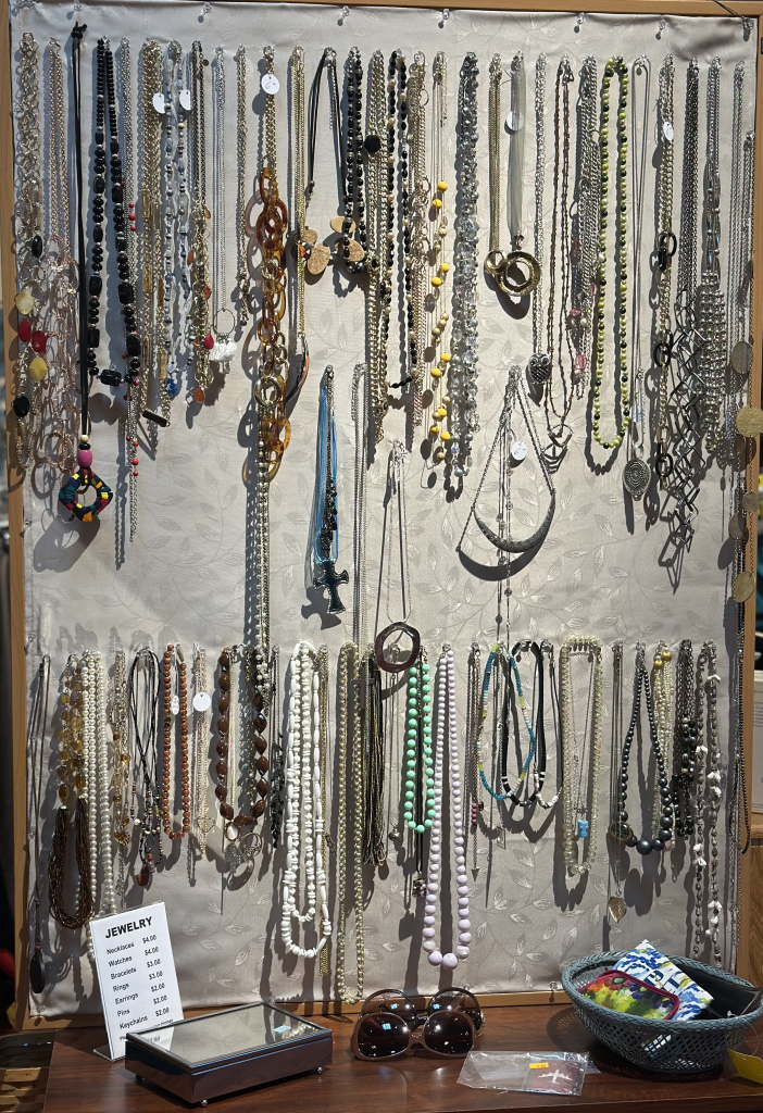 Display of assorted necklaces hanging on a wall-mounted board, with various beads, chains, and pendants. A small table below holds sunglasses, a basket with items, and a price card labeled ‘Jewelry.’