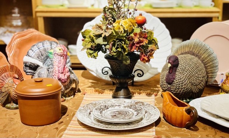 Thanksgiving‑themed table display featuring ceramic turkey figurines, fall floral centerpiece, stacked patterned plates, an orange gravy boat, and a lidded pot arranged on a decorative tablecloth.