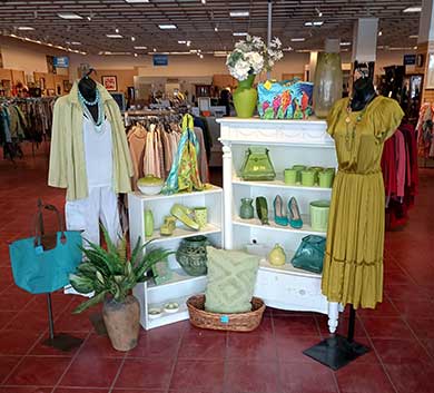 Boutique retail display featuring coordinated spring apparel in light green and neutral tones, including dresses, jackets, handbags, shoes, and accessories arranged around white shelving inside a clothing store.
