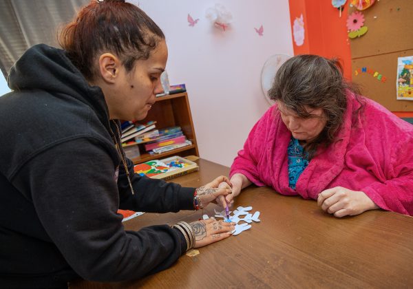 Two individuals are seated at a wooden table decorating a paper snowflake together. The person on the left is wearing a black hoodie, while the person on the right is wearing a pink robe. The background includes shelves with books and colorful decorations on the walls.