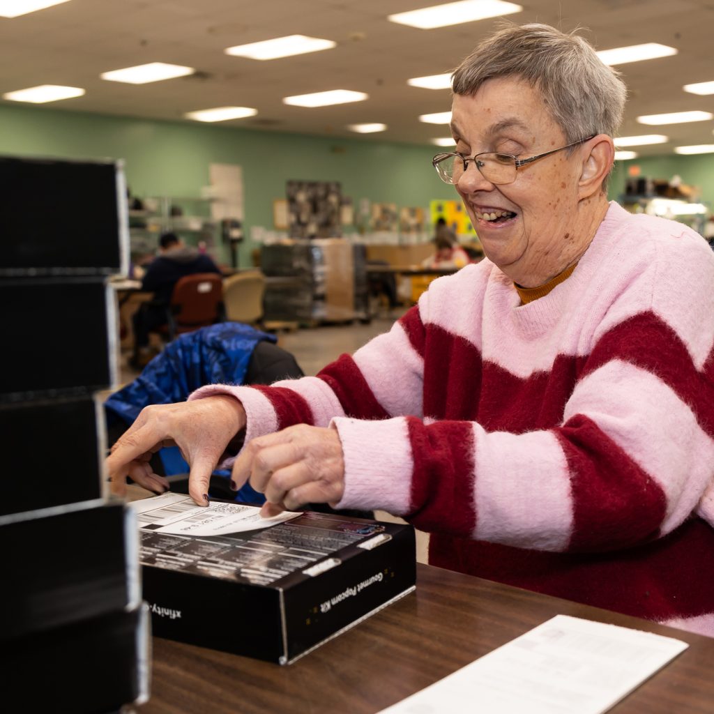 A person wearing a pink and red striped sweater is sitting at a table in a large room with fluorescent lighting. The person is affixing white labels to black boxes. Other people are working in the background, and the room has green walls and various office furniture.