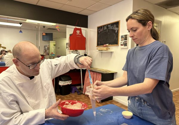 A person in a white chef's coat and another person in a blue shirt are working together in a kitchen. The person in the chef's coat is holding a red bowl with batter, while the other person is using a piping bag to fill cupcake liners. The background shows kitchen equipment, an apron hanging on the wall, and a chalkboard with "Cake of the Day" written on it.