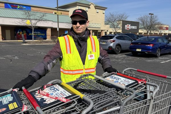 A person wearing a yellow reflective safety vest and a black cap is pushing a row of shopping carts in a parking lot. The person is smiling. In the background, there are parked cars and a storefront with visible signage. The sky is clear and blue.